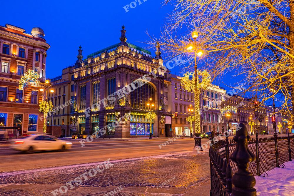 Фотоснимок передаёт атмосферу уютного зимнего вечера: подсвеченные фасады зданий, огни фонарей, украшенные деревья и отражения света на дороге. Это изображение подчёркивает красоту архитектуры Петербурга, его романтичный облик и праздничное настроение. Картинка может использоваться как иллюстрация или слайд, посвящённые зимнему городу, путешествиям и городской жизни Санкт-Петербурга.